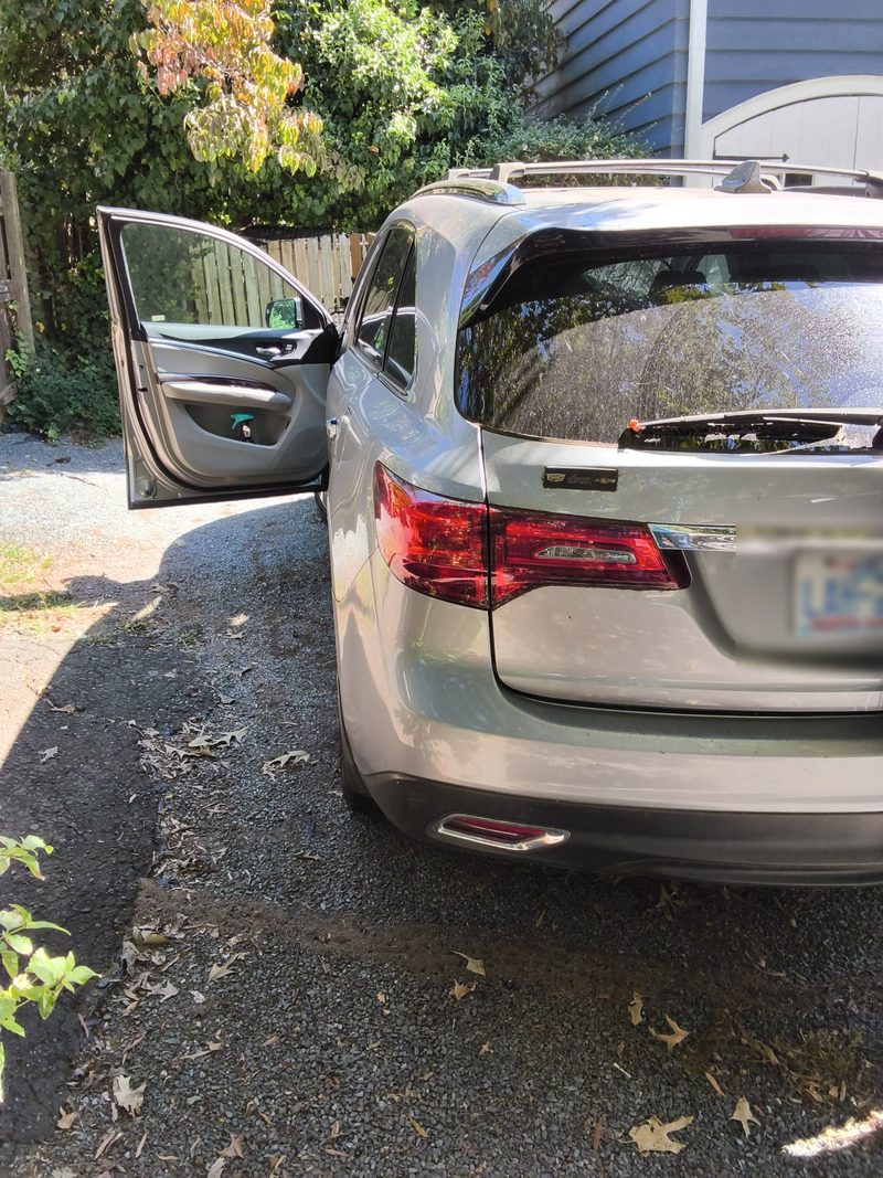 The back of the silver Acura MDX on a gravel driveway with the driver door open and the plate blurred