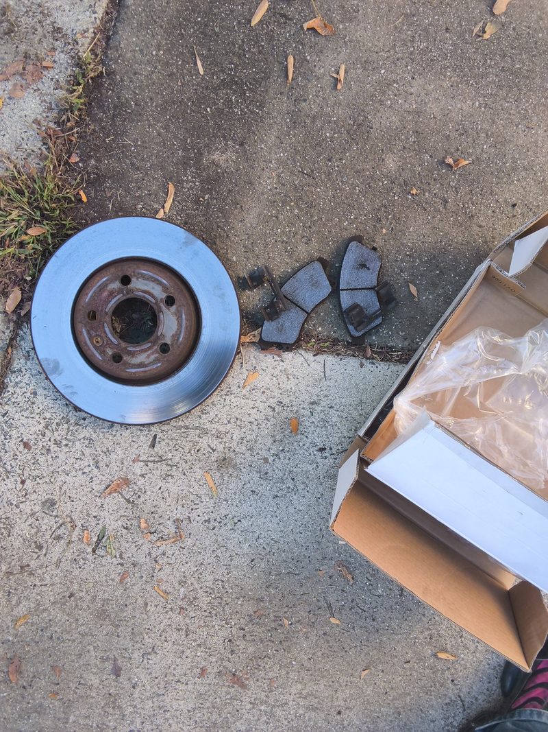 The old front rotor and worn pads from the Cruze on concrete beside the new parts box