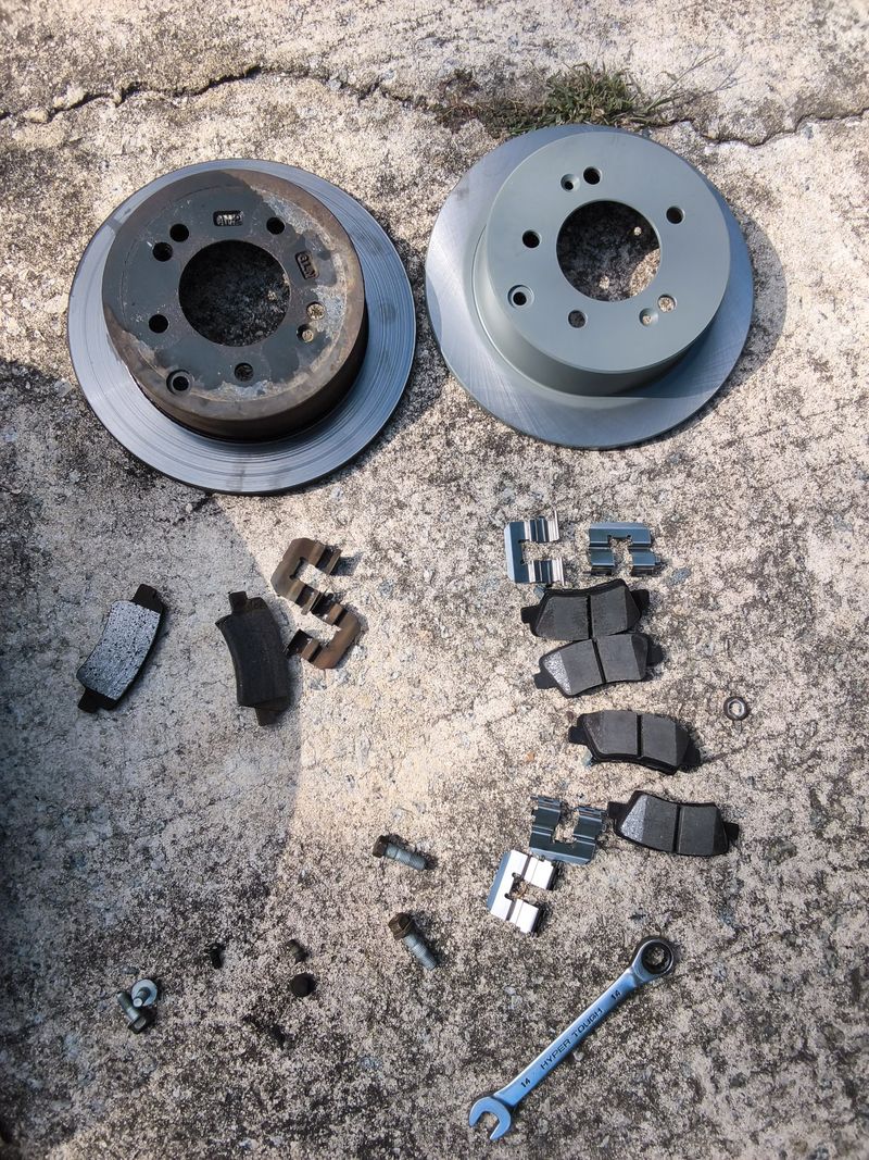Worn rear brake rotors and pads beside the new set on a concrete driveway