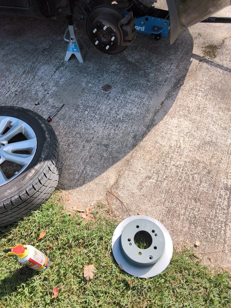 A new brake rotor on the grass beside the Kia while the car sits on a jack stand