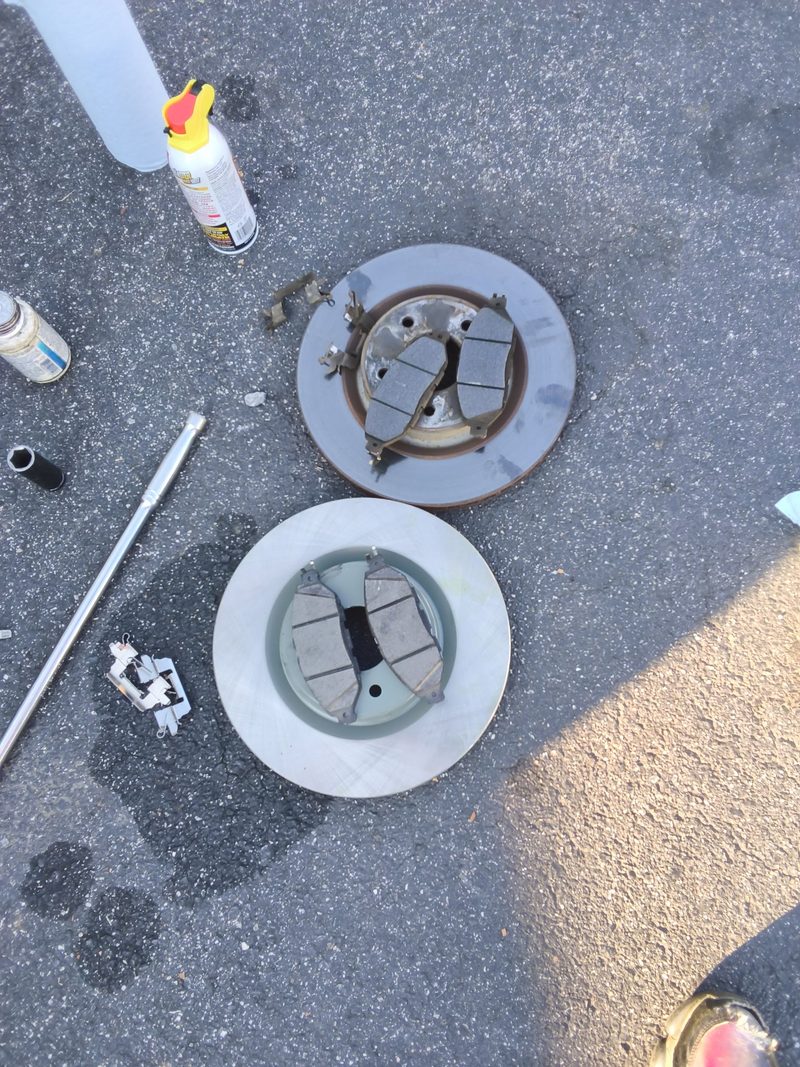 A rusty old rotor with old pads beside a clean new rotor with new pads on the asphalt