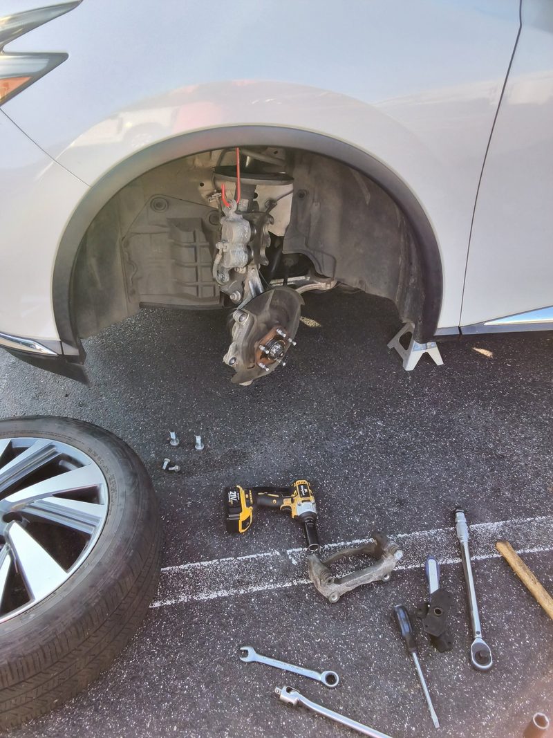 The front corner of the white Murano on a stand with the wheel off and tools on the asphalt