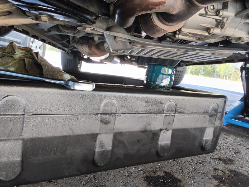 A black oil drain pan under the Honda Pilot with the jack and the underside of the engine above it